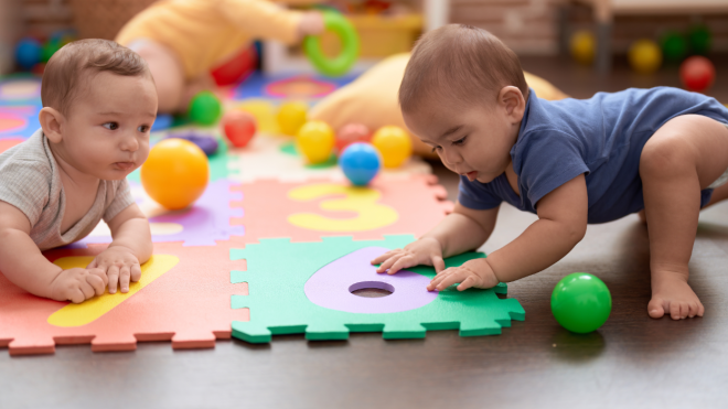 Two young infants play together on a foam mat with number cutouts and colourful balls