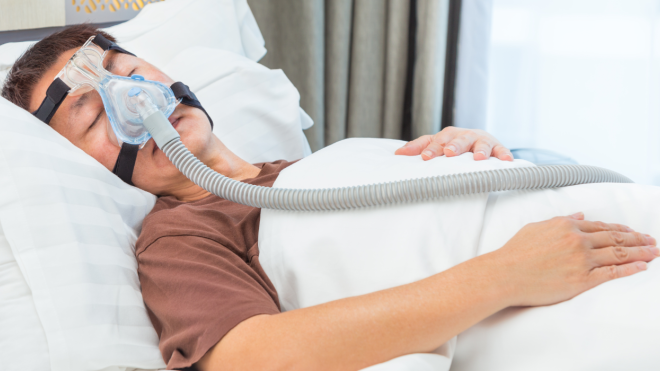 A man in a brown shirt, sleeping on a white pillow, with white covers wears a CPAP mask.