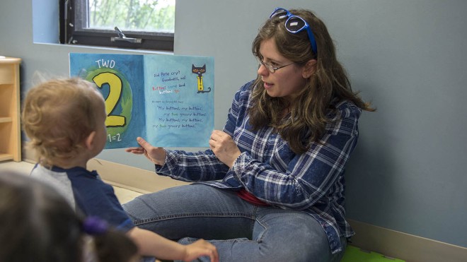 An individual sits on the floor of a classroom at the Simcoe/Norfolk Regional Campus, reading a book to a small group of children as part of the Early Childhood Education program; the book is open to a page with the number "2" and a cartoon cat.