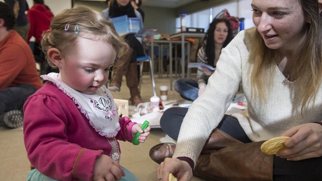 Inside a classroom at Fanshawe College's School of Community Studies, a child in a pink sweater and turquoise pants sits on the floor, holding a green toy. A person in a cream-colored sweater and brown boots sits cross-legged nearby, holding a yellow toy. Other people are seated in the background.