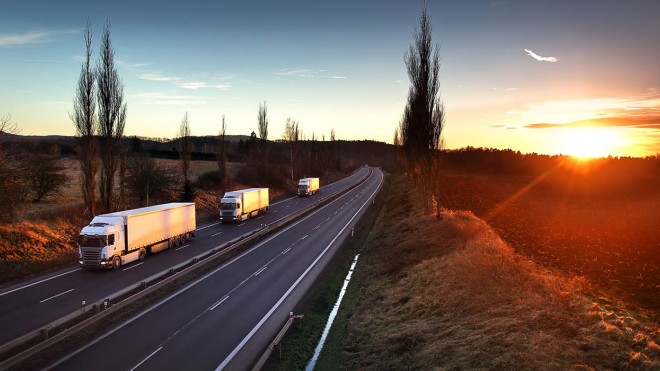 The image shows a multi-lane highway with three large trucks driving on it, with a field and trees on either side, and a bright sunset in the distance.