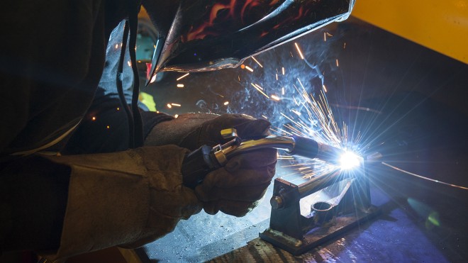 A person wearing a flame-patterned welding mask and protective gloves welds metal, creating bright sparks and smoke.