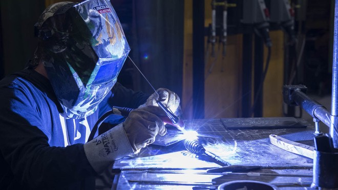 A person wearing a welding mask and gloves is welding metal on a table, with bright sparks flying.