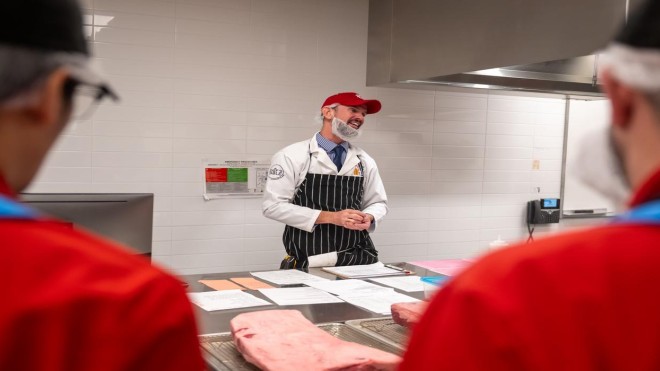 At the London Downtown Campus, in a room with white tiled walls and stainless steel counters, a person in a white lab coat, red baseball cap, and black and white striped apron stands behind a counter with papers and a large cut of meat, while two people in red jackets stand in the foreground.
