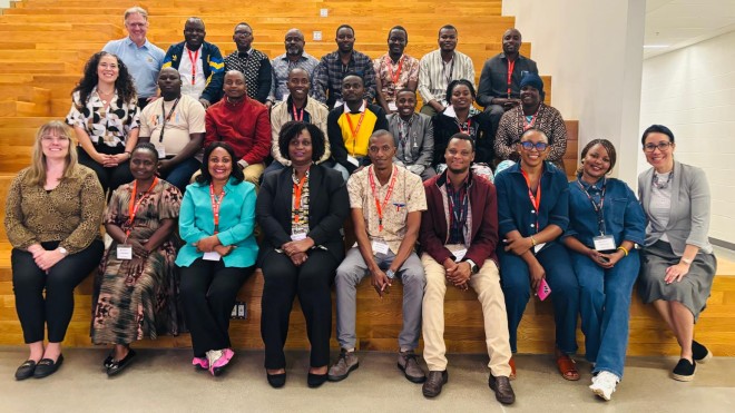 A large group of Tanzanian educators and Fanshawe Global representatives pose together on tiered wooden seating in a modern indoor space.