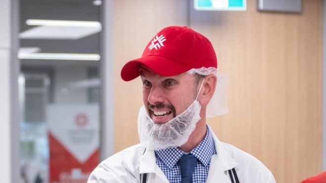 At the London Downtown Campus, Troy Spicer wearing a white lab coat, a red baseball cap, and a hair net gestures over a white cutting board with various cuts of meat, while another person wearing a black baseball cap and a red shirt stands in the background.