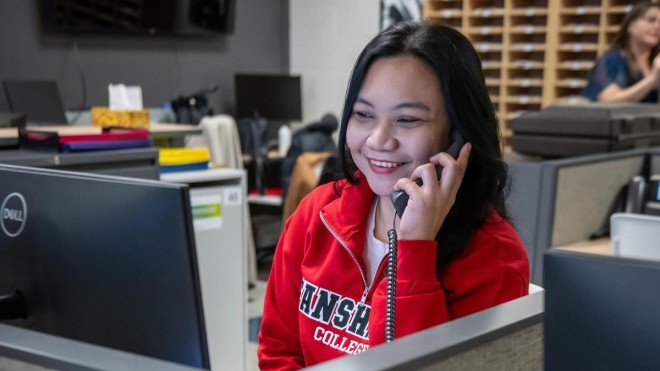 In a Fanshawe College London Campus Office Administration classroom, a person in a red Fanshawe College jacket smiles while talking on a telephone in a cubicle.