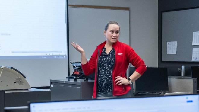 In a classroom at Fanshawe College's London Campus, a person stands near a projector screen displaying a spreadsheet, gesturing with one hand while the other rests on their hip. In the foreground, another person sits at a desk with a computer monitor displaying a Microsoft update screen, a keyboard, a mouse, and a desk phone. The desks are separated by cubicle walls. Another person sits at a desk in the background, facing away from the camera. A whiteboard, clock, and bulletin board are visible on the walls
