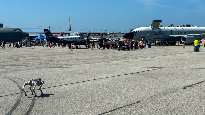 Robot dog on an airport tarmac during an airshow, with aircraft and visitors in the background.