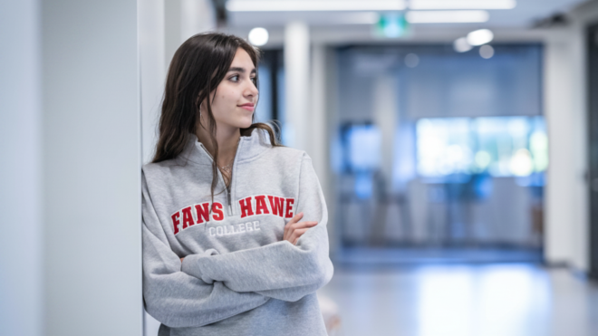A person wearing a grey sweater with red lettering leans against a white wall in a hallway.