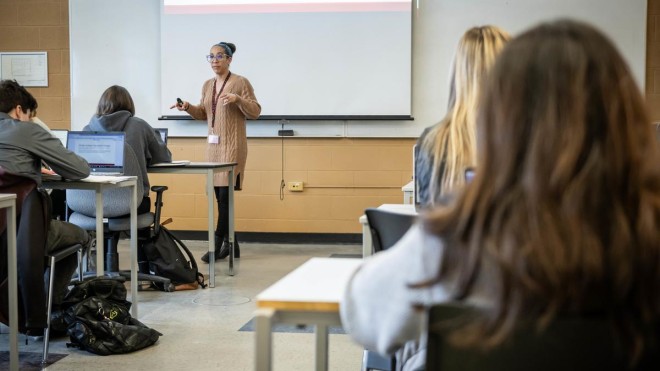 Inside a classroom at Fanshawe College's London Campus, a person stands near a projection screen, gesturing with a remote in their hand, while others sit at desks with laptops.