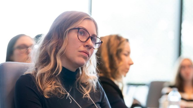 At the London Campus of Fanshawe College, a person with glasses and long wavy hair sits in a blue chair, wearing a black turtleneck and grey pants, with a tan jacket draped over the side of the chair.