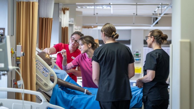In a simulated hospital room, two individuals in pink and red scrubs examine a medical mannequin lying in a blue-sheeted hospital bed, while two others in black scrubs observe.