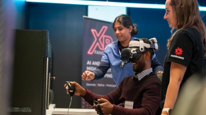 A person wearing a virtual reality headset and holding handheld controllers sits at a desk while two others observe nearby.