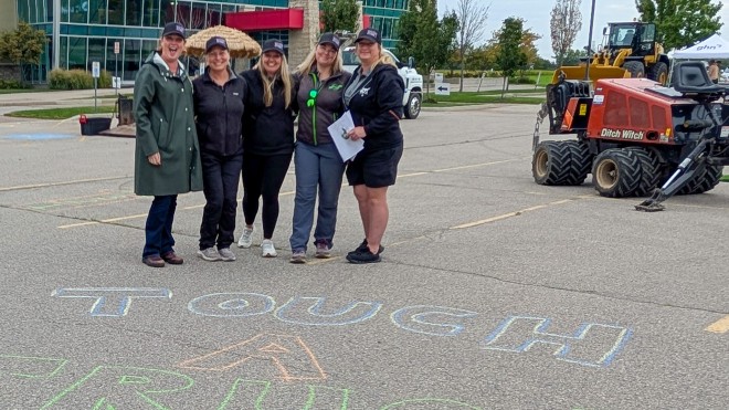 Five women stand in a line smiling behind "Touch a Truck" written in chalk on the concrete.