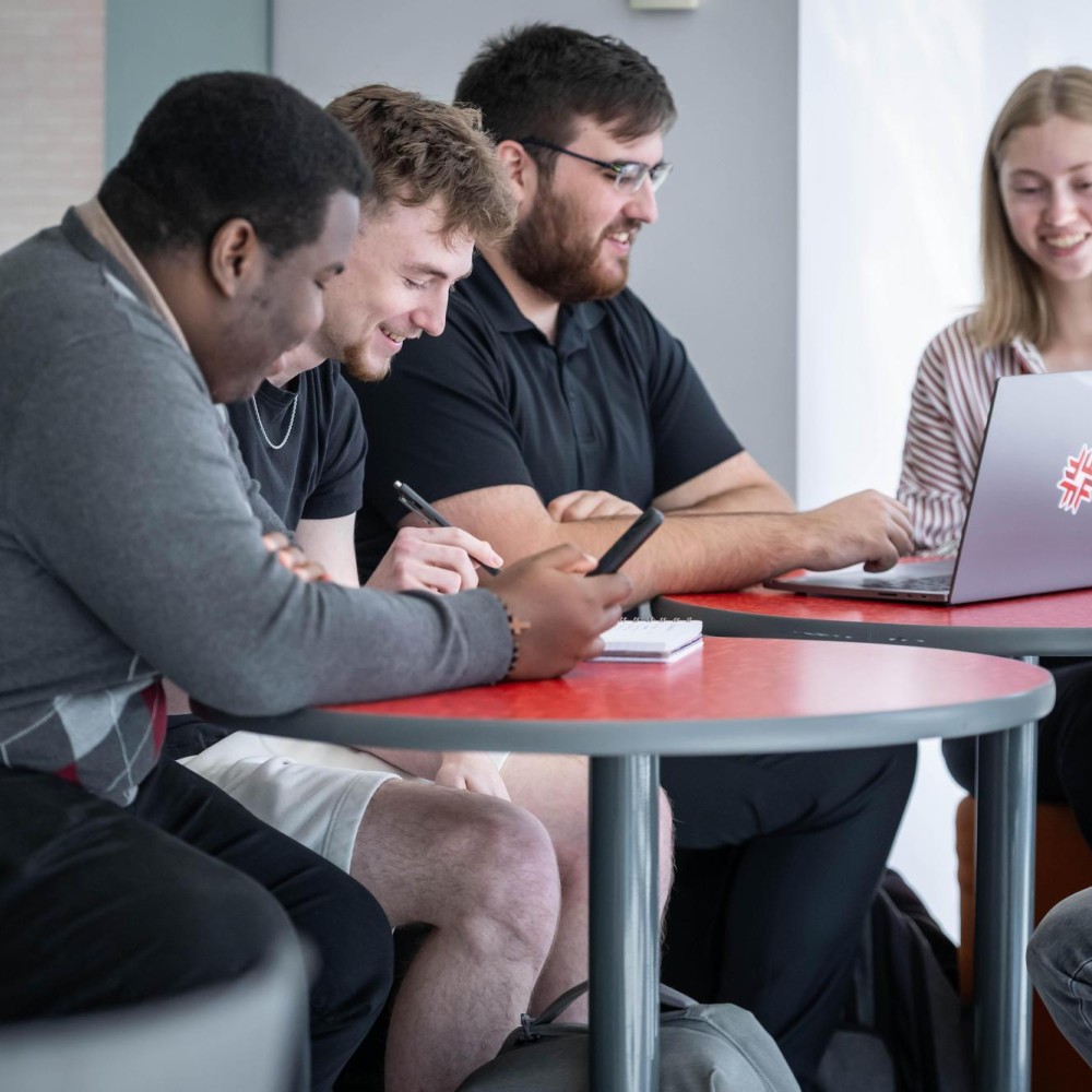 A group collaborates at a red table with a laptop bearing the Fanshawe College logo, inside the London Downtown Campus.