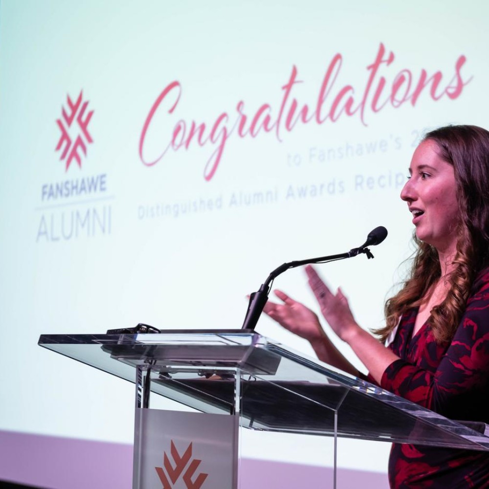 At the Fanshawe College Distinguished Alumni Awards event, a person stands at a clear podium with a microphone, in front of a screen that reads "Congratulations to Fanshawe's Distinguished Alumni Awards Recipients."