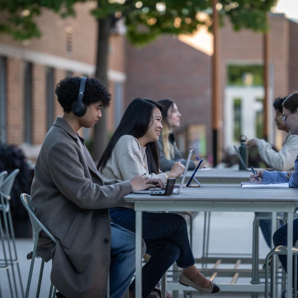 Students work at outdoor tables on Fanshawe College's London Campus, using laptops, tablets, and notebooks.