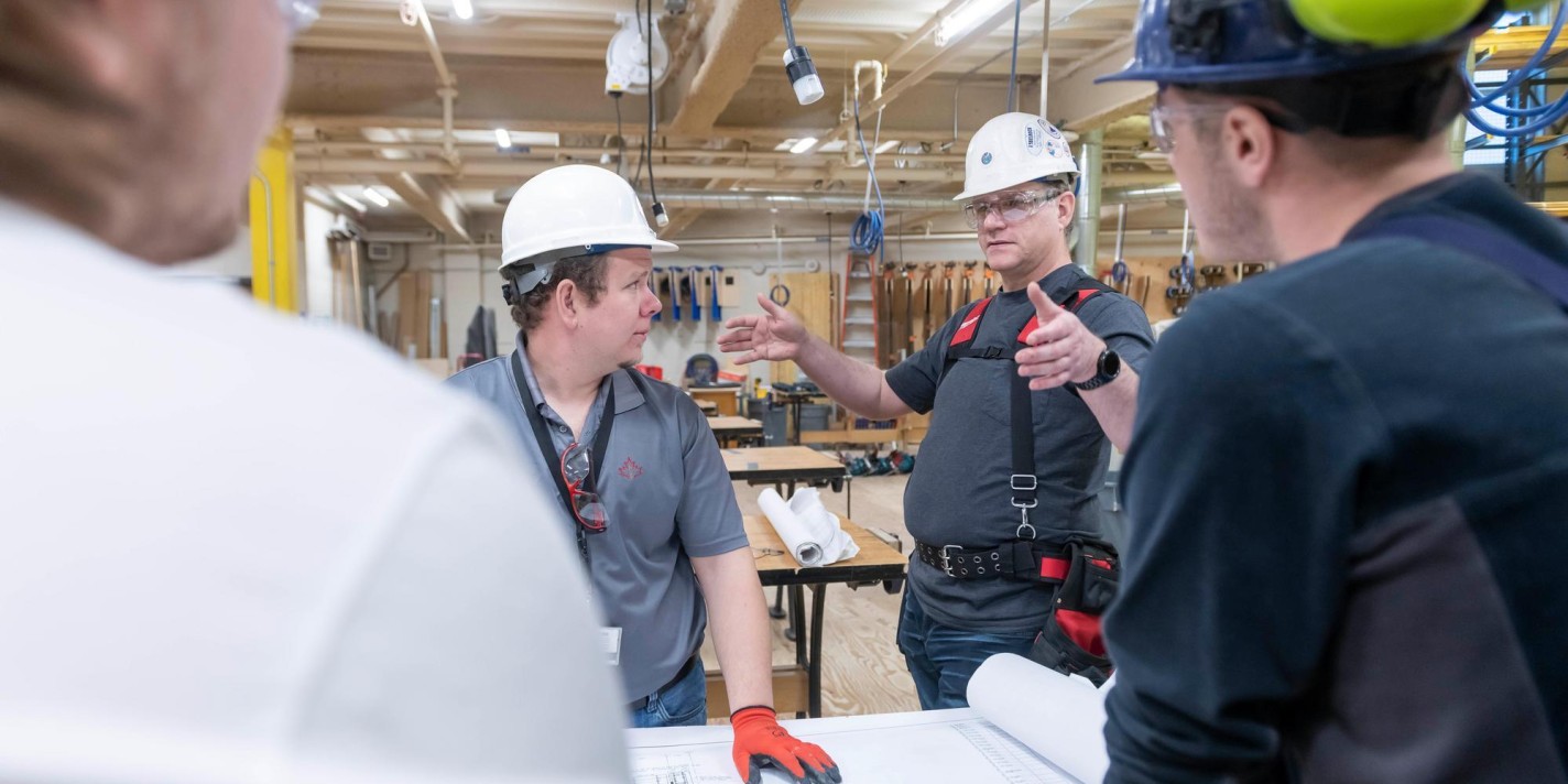In a Fanshawe College workshop, individuals wearing hard hats and safety glasses examine a large blueprint spread across a table.