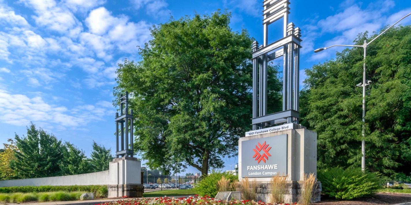 A sign with a red and white logo sits on a concrete base, framed by a metal structure, with a flower bed of red and white flowers in the foreground.