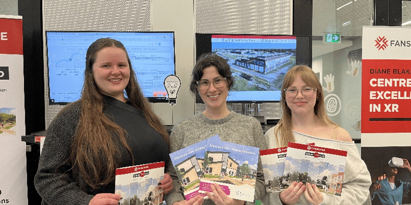 Three people smile while holding brochures in a tech-themed room with posters and screens in the background. The mood is welcoming and enthusiastic.