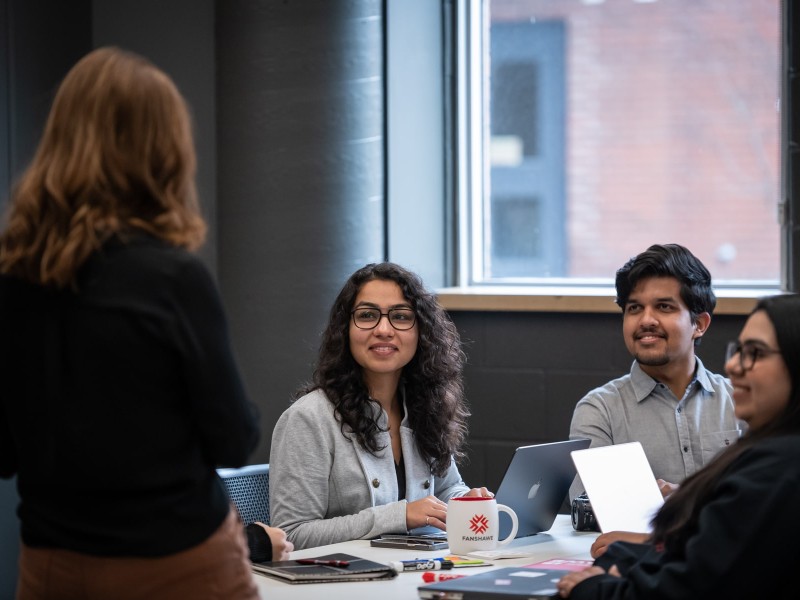 students sit at table with laptops, listening to presenter