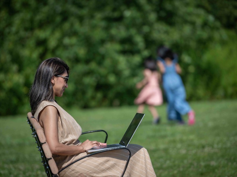 student sits outside on laptop while children play in background