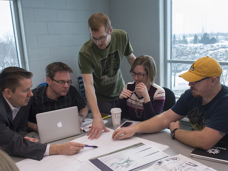 At the London Campus of Fanshawe College, a group gathers around a table covered with architectural drawings, a laptop, and notebooks, discussing plans with a large window overlooking a snowy parking lot in the background.