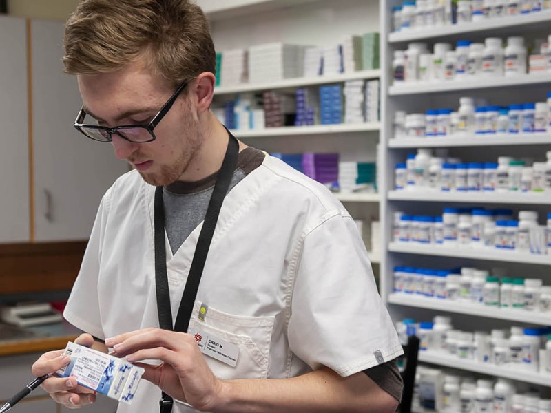 Inside the School of Health Sciences at Fanshawe College's London Campus, a person wearing glasses and a white lab coat holds a blister pack of medication and a pen, standing in front of shelves stocked with numerous bottles of pills.