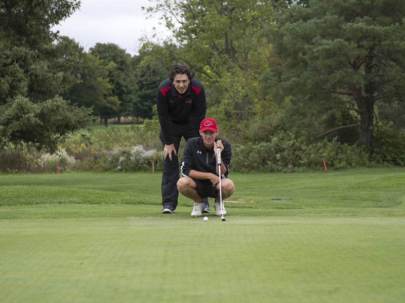 On a green golf course, one person crouches with a putter near a golf ball, while another person stands behind them, observing. A golf bag sits nearby, and trees line the background.