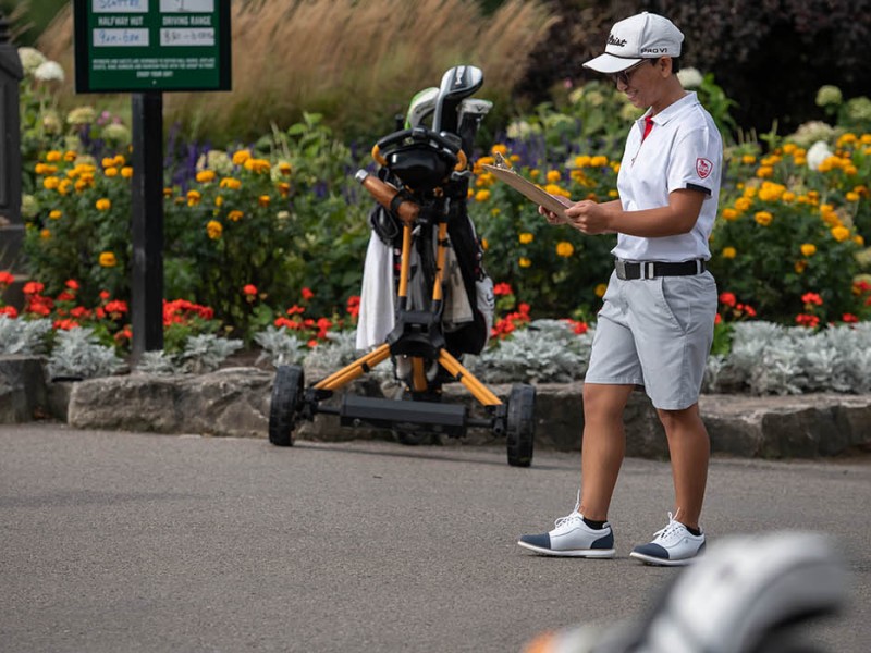 At a golf course, a person in a white polo shirt, grey shorts, and a white cap stands on a paved path, holding a clipboard, with golf bags and carts nearby, and a course status sign visible behind a flower bed.