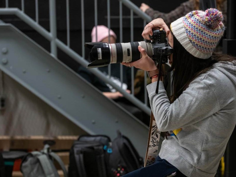 At the London Campus of Fanshawe College, someone wearing a colourful toque and grey hoodie sits on a wooden box, holding a camera with a long lens up to their eye, with a grey staircase and other people in the background.