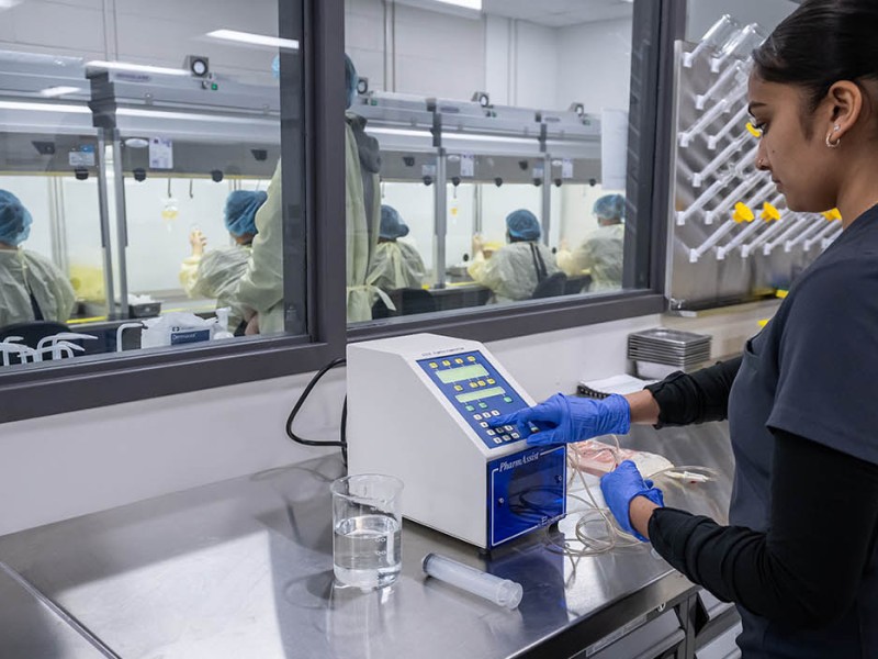 At the School of Health Sciences of Fanshawe College at the London Campus, a person wearing a grey uniform with a Fanshawe College Community Pharmacy Assistant Student patch is pressing a button on a keypad.