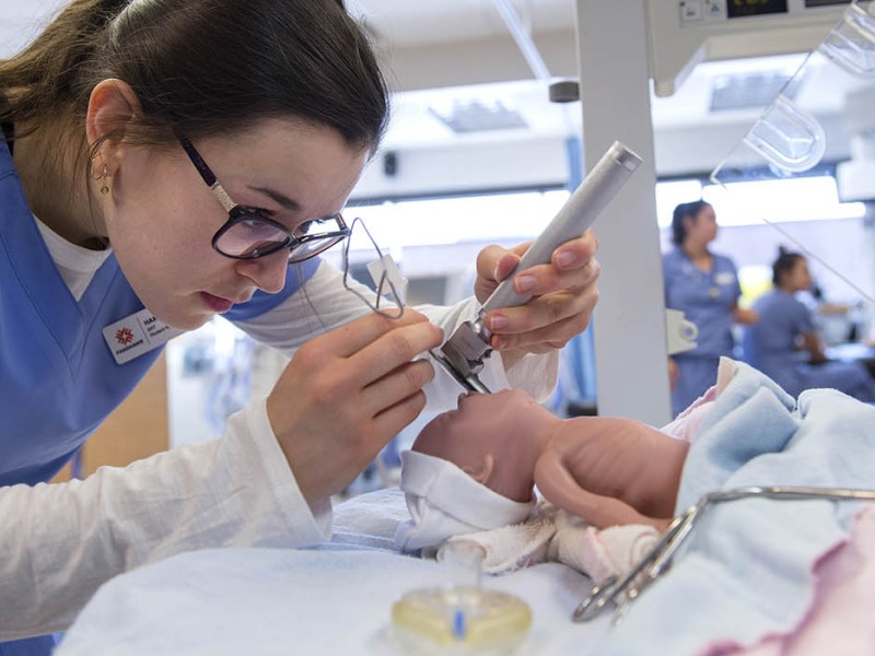 In the School of Health Sciences at Fanshawe College's London Campus, a person wearing a blue scrub top and glasses uses medical instruments to examine a baby mannequin lying on a bed with blue and pink blankets.