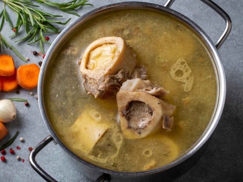 A steel pot of golden liquid, bones and other ingredients on a counter with some garlic, onion, carrots and rosemary. 