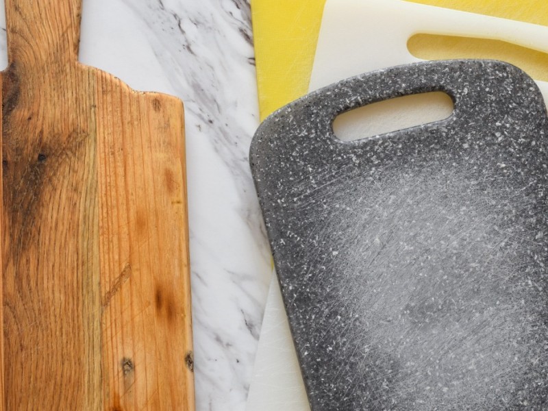 One wooden cutting board beside a stack of plastic cutting boards on a marble counter.
