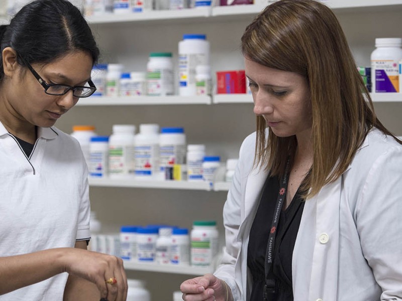 At the School of Health Sciences of Fanshawe College at the London Campus, two people stand in front of shelves stocked with various medications; one person holds a small container, while the other holds a few pills.