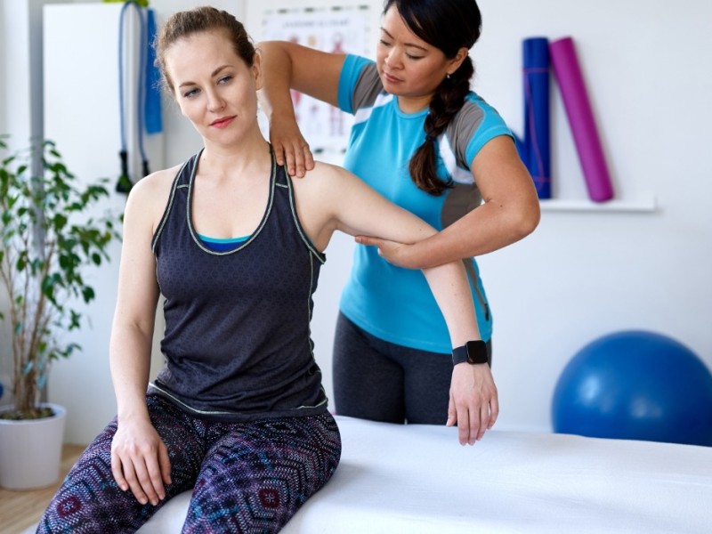 A woman sits on a therapy table, as a physiotherapy practitioner works on her shoulder.  A blue yoga ball is blurred in the background along with an indoor plant, yoga mats and tension cords. 