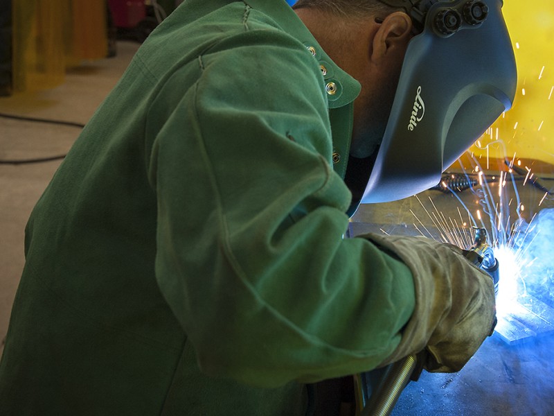 A person wearing a green jacket, gloves, and a welding mask is welding metal, creating bright sparks and blue light. The person is positioned in front of a yellow wall, with welding equipment and gas tanks visible in the background.