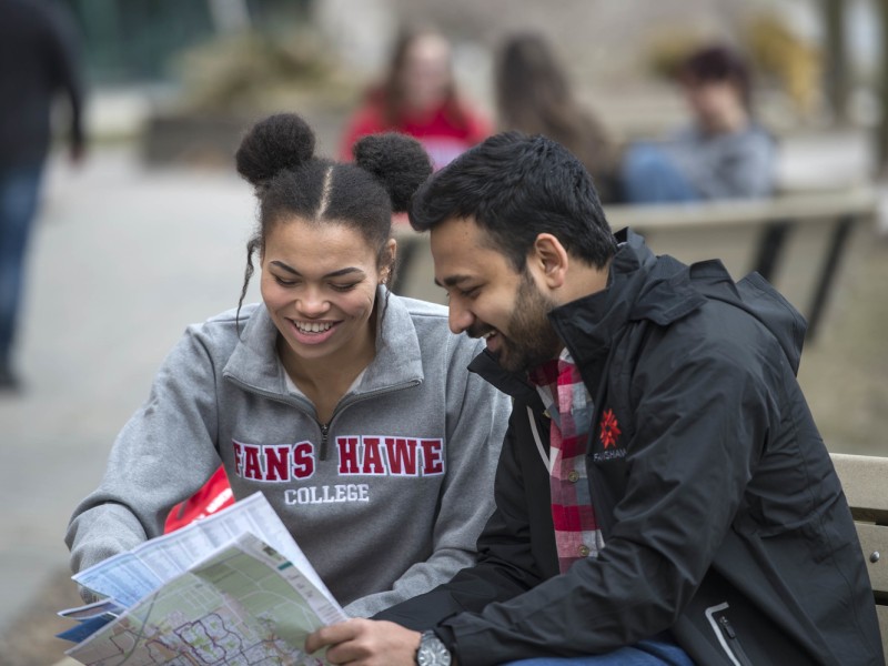 Two people wearing Fanshawe College branded clothing look at a map while sitting on a bench at Fanshawe College's London Campus.