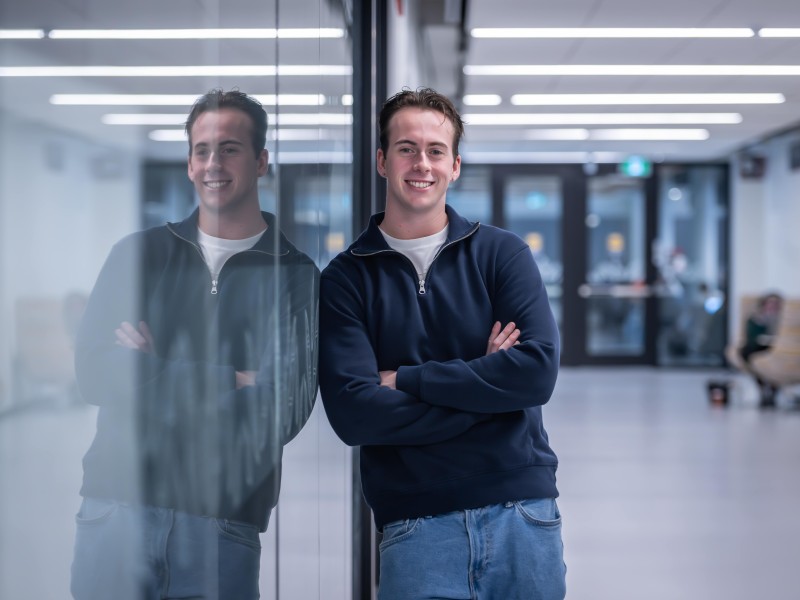 A person in a navy sweater and jeans stands with arms crossed, leaning against a glass wall that reflects their image.