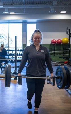 At the Fanshawe College London Campus, a person in the Fitness and Health Promotion program carries a barbell with weights in a gym setting, with other exercise equipment and people visible in the background.