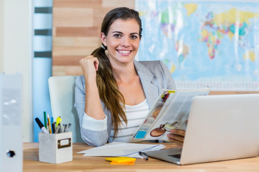 Smiling woman in a gray blazer with a ponytail sits at a desk with a laptop, holding a magazine. A world map and stationery are visible in the background.