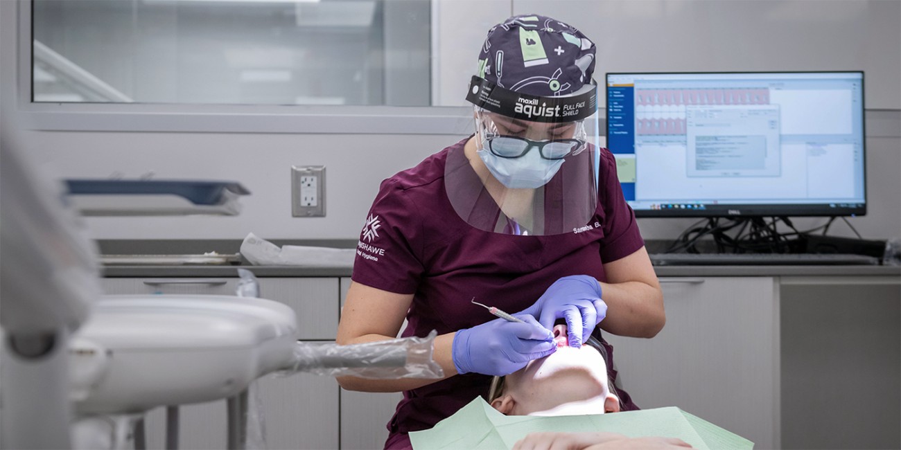 A dental hygiene student wearing a face mask, gloves, and scrubs is using a dental tool to examine a patient lying in a medical chair. A computer monitor displays dental information in the background.