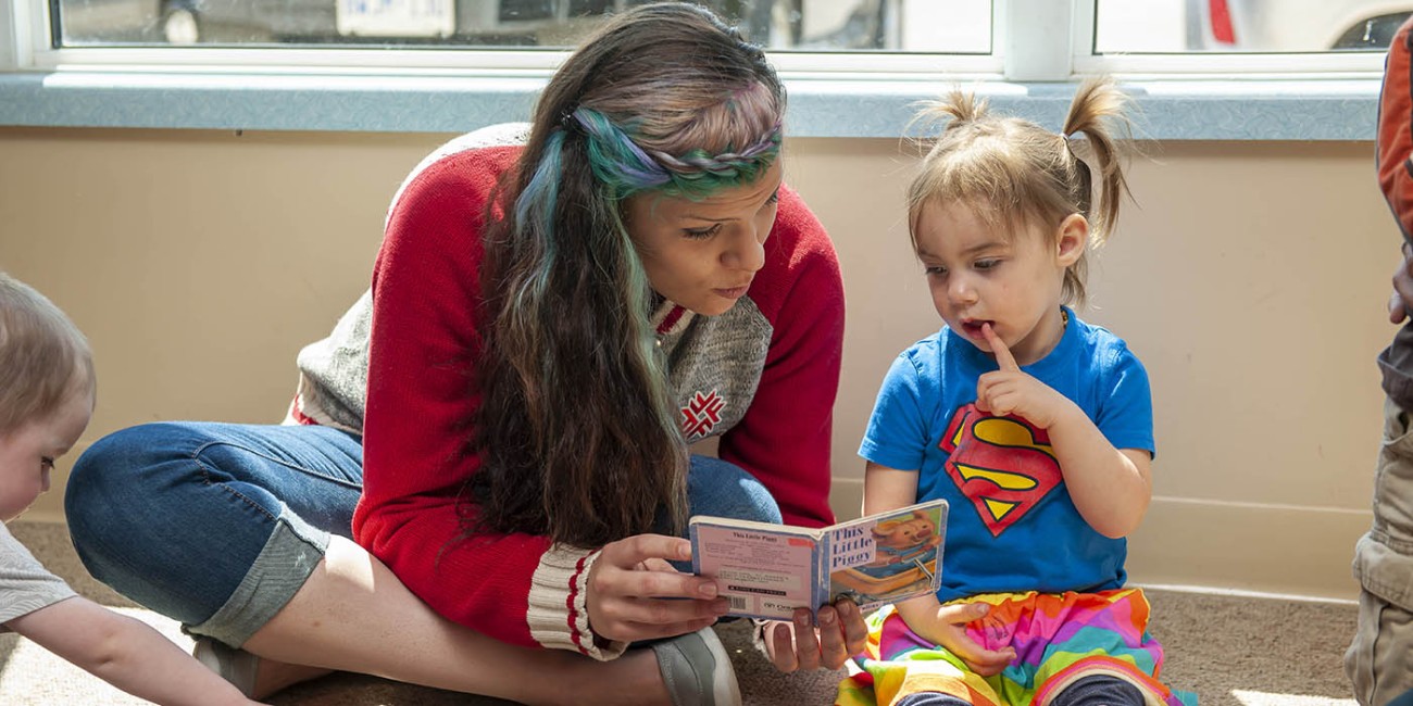 At the St. Thomas/Elgin Regional Campus of Fanshawe College, an individual sits cross-legged on the floor reading "This Little Piggy" to a child wearing a Superman t-shirt and rainbow-striped pants, while another child plays with a wooden train set nearby.
