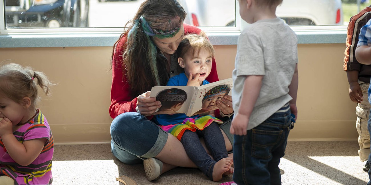 At the Fanshawe College St. Thomas/Elgin Regional Campus, an individual with long brown hair and blue and green highlights sits cross-legged on the floor, reading a book titled "Faces of Tomorrow" to a child in a blue shirt and rainbow skirt, while other children stand nearby. The scene is set 