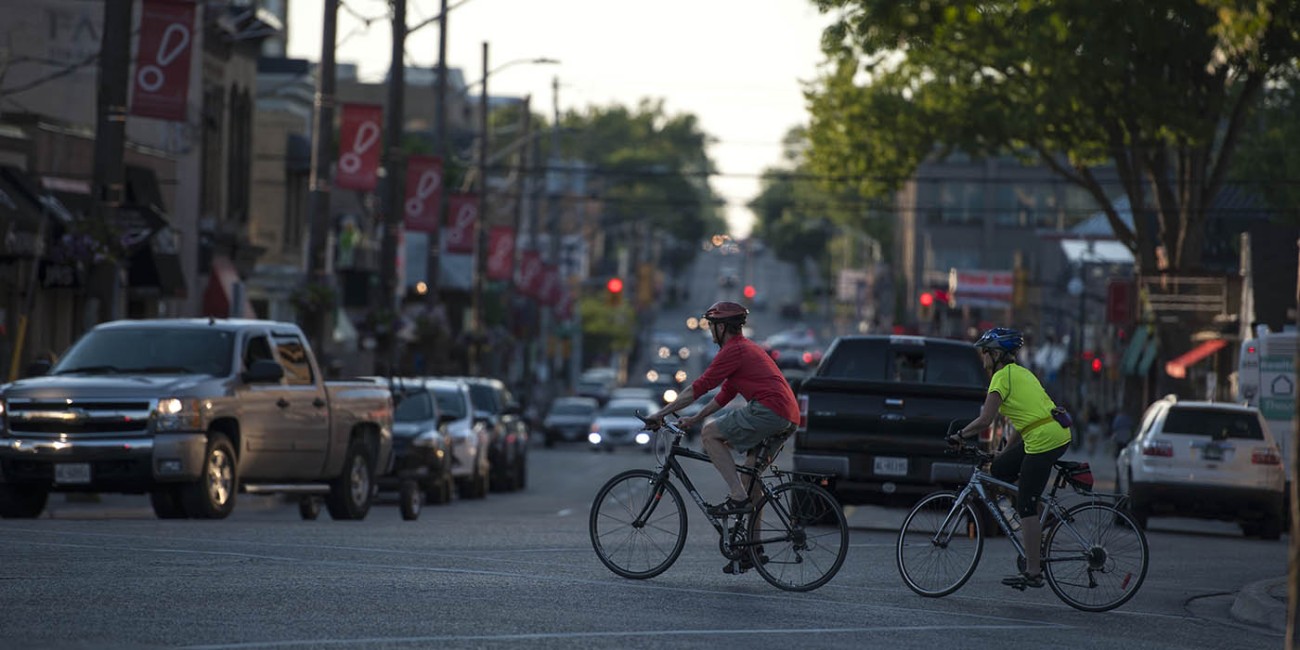 Two cyclists ride across a city street, sharing the road with cars, likely for transportation or recreation.