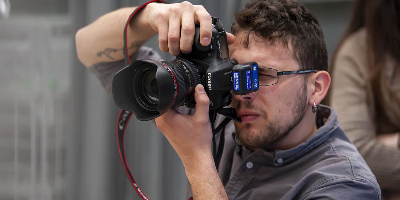At the Fanshawe College London Campus, someone holds a Canon camera up to their face, looking through the viewfinder, with a flash attachment on the camera.