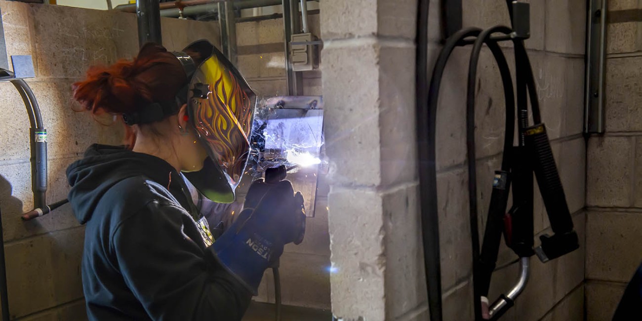 At the Tiverton location of the Huron-Bruce Regional Sites of Fanshawe College, a person wearing a flame-designed welding mask and protective gloves is welding metal in a welding bay, as part of the Welding Techniques program.