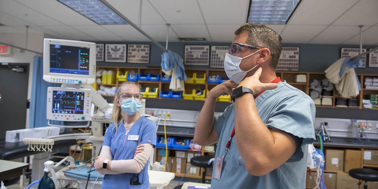In the School of Health Sciences at Fanshawe College's London Campus, four people in blue scrubs and face masks are gathered around medical equipment, including a ventilator and monitors displaying medical data, with a mannequin lying on a table in the foreground.